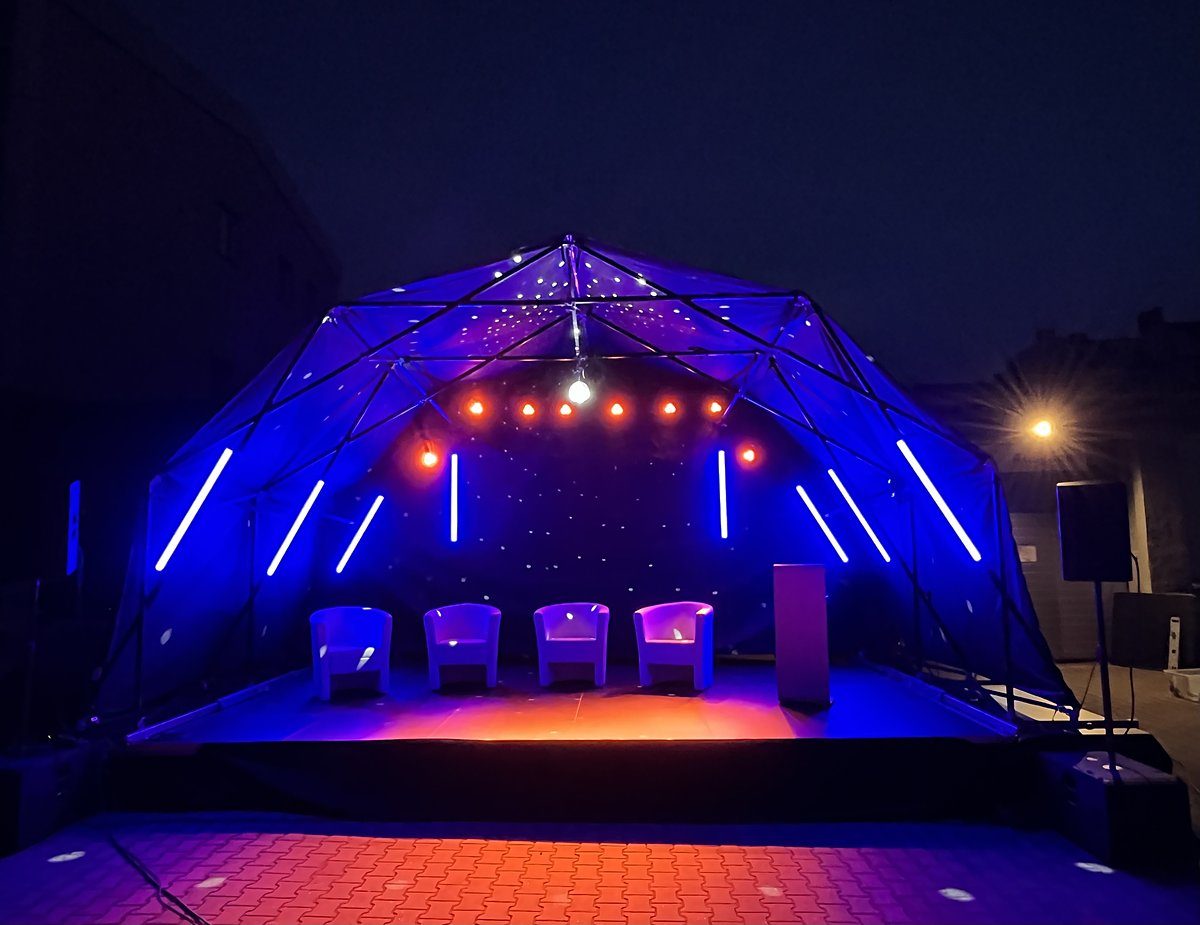 Open-Air-Bühne bei Nacht Open-Air-Bühne mit Rundbogendach bei Nacht, beleuchtet mit blauen Astera Titan Tubes und warmen Spots